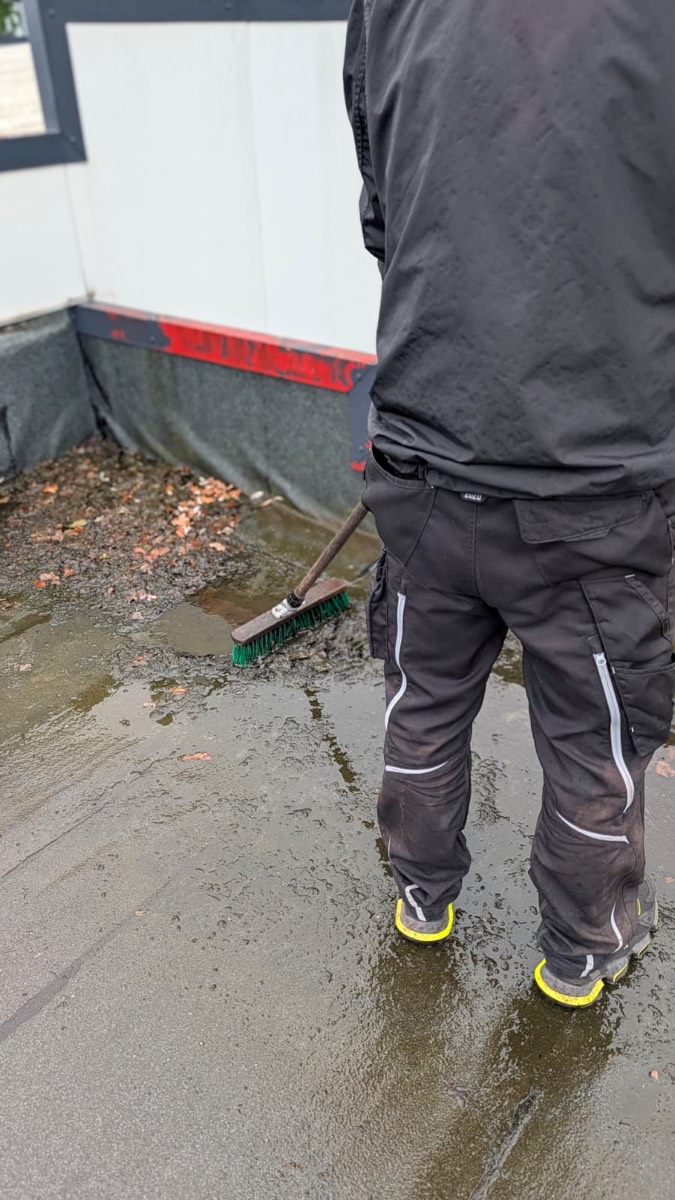 Rückenansicht eines Mitarbeiters, der Laub und Schlamm an der rot abgesetzten Attika zusammenkehrt