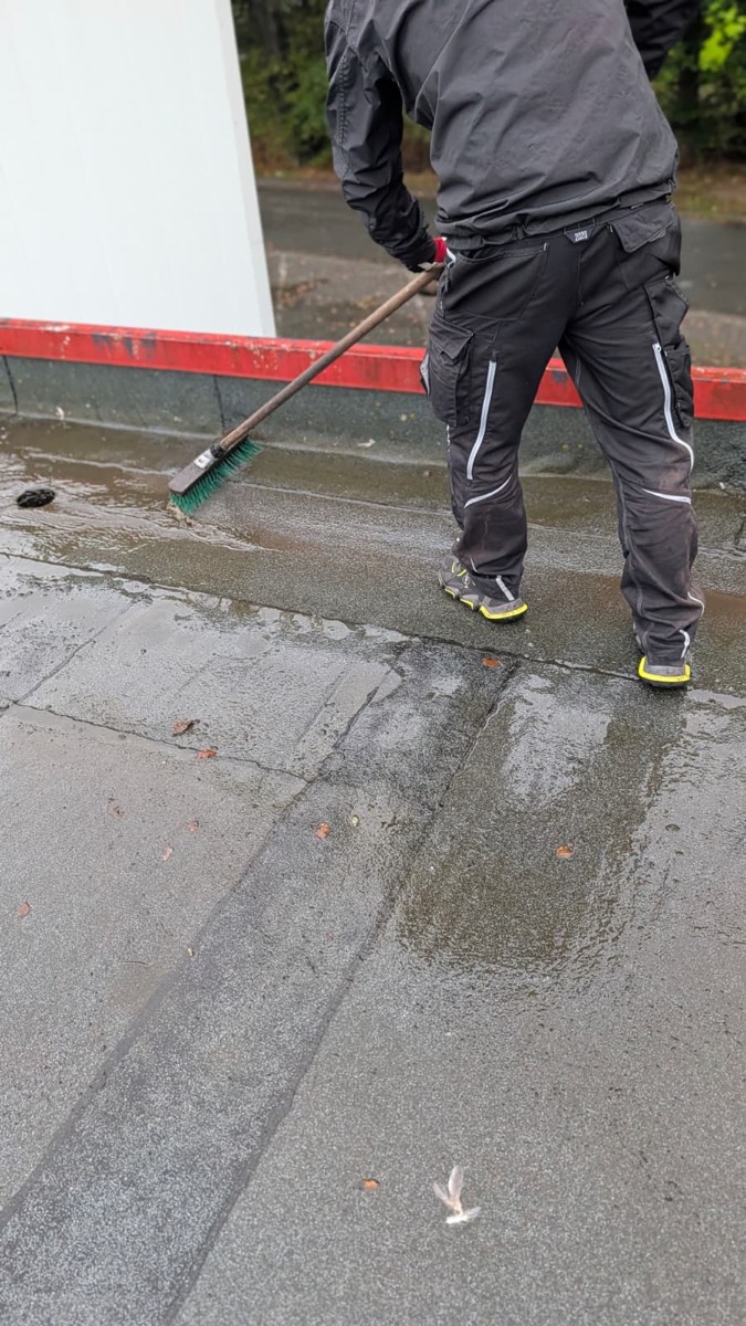 Ein Mitarbeiter schiebt mit dem Besen Regenwasser über die Dachbahn zum Ablauf an der Attika