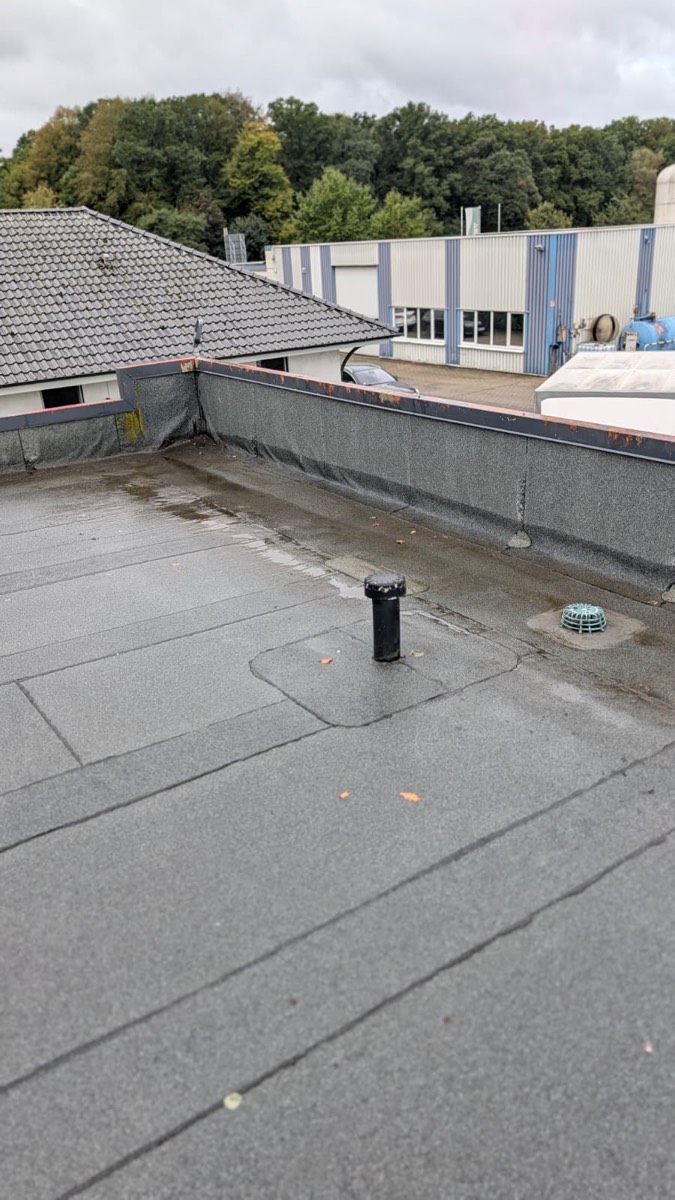 Flachdachfläche nach dem Abfließen des Wassers, der Einlaufkorb an der Attika liegt frei, dahinter Bäume und eine Halle