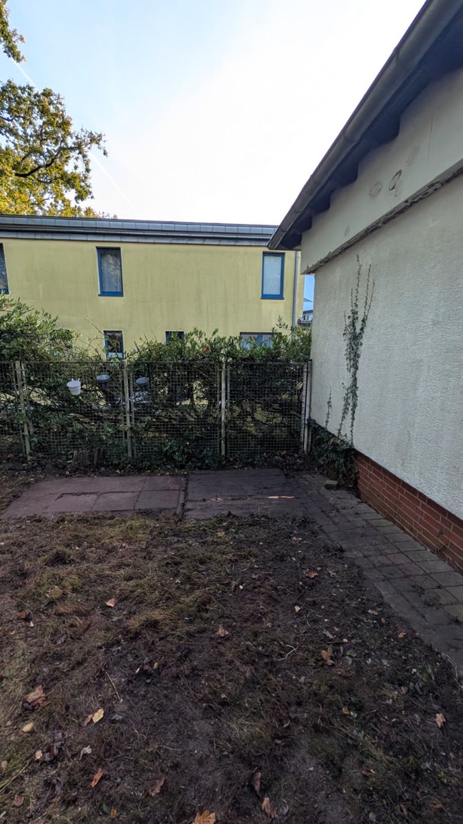 Die Ecke ohne Baum: offener Erdboden, freier Blick auf Gitterzaun, Plattenweg und das gelbe Nachbarhaus